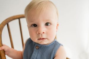 A small child wearing a blue knit outfit sits on a wooden chair in a minimalist white setting.