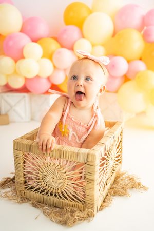 Joyful baby girl in pink romper sitting in wicker basket, surrounded by pink and yellow balloons.