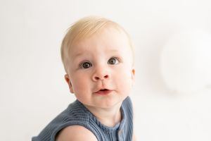 Baby wearing blue denim clothing smiles at the camera with bright eyes and rosy cheeks.