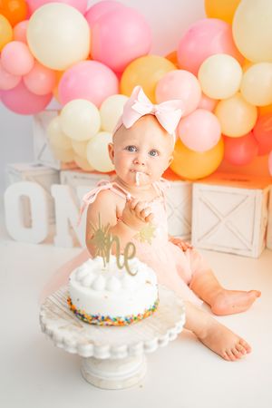 Baby girl in pink tutu celebrates first birthday smash cake session with colorful balloon backdrop.