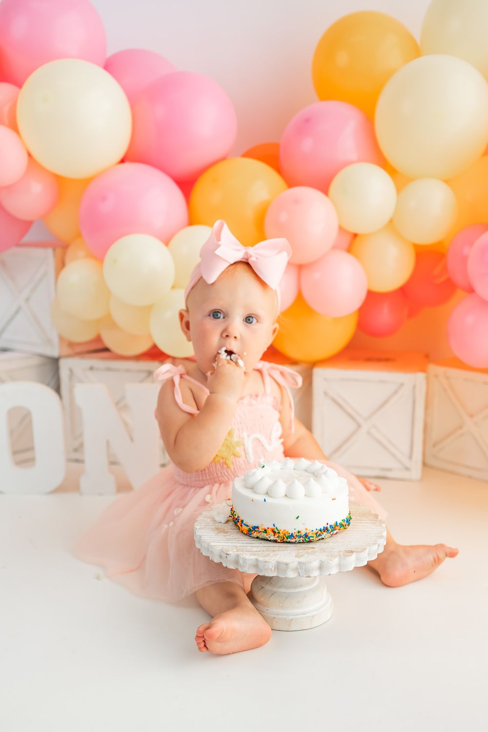 Baby girl in pink tutu eating cake at first birthday party with colorful balloon backdrop.