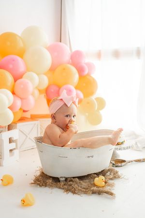 Baby girl in pink bow sits in vintage white tub with rubber ducks and colorful balloon backdrop.