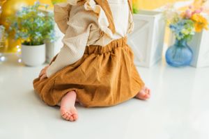 Close-up of tiny bare feet under brown dress and cream sweater on white floor.