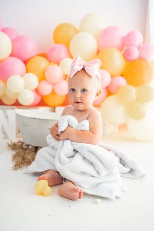 Baby girl wrapped in white blanket with pink bow headband sits before colorful balloon backdrop.