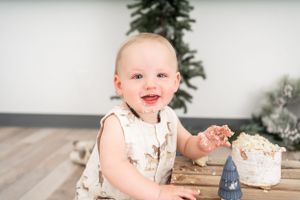 Baby in white dress sitting with cake and holiday decorations in background.