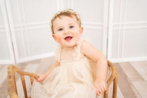 Baby in cream dress smiles joyfully while sitting in antique wooden chair against white background.