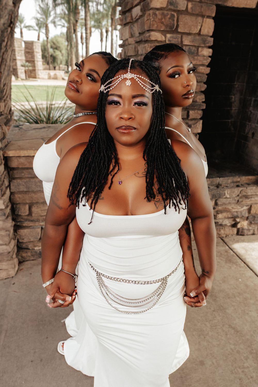 Three women in white dresses pose with decorative head chains under brick archway.