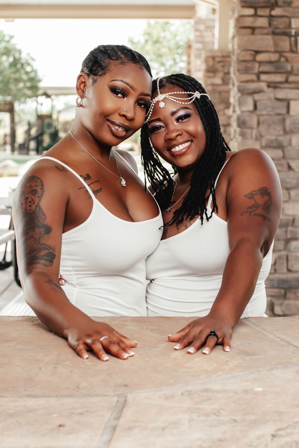 Two women in white tank tops pose together under brick archway with decorative headpieces.