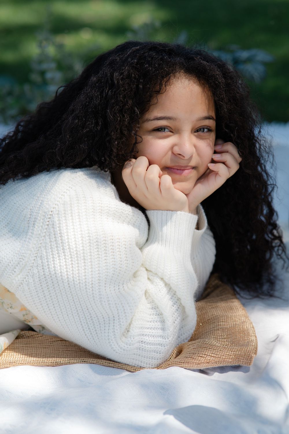 Person with curly dark hair wearing a white knit sweater lays on a blanket outdoors while smiling and resting on their hands.