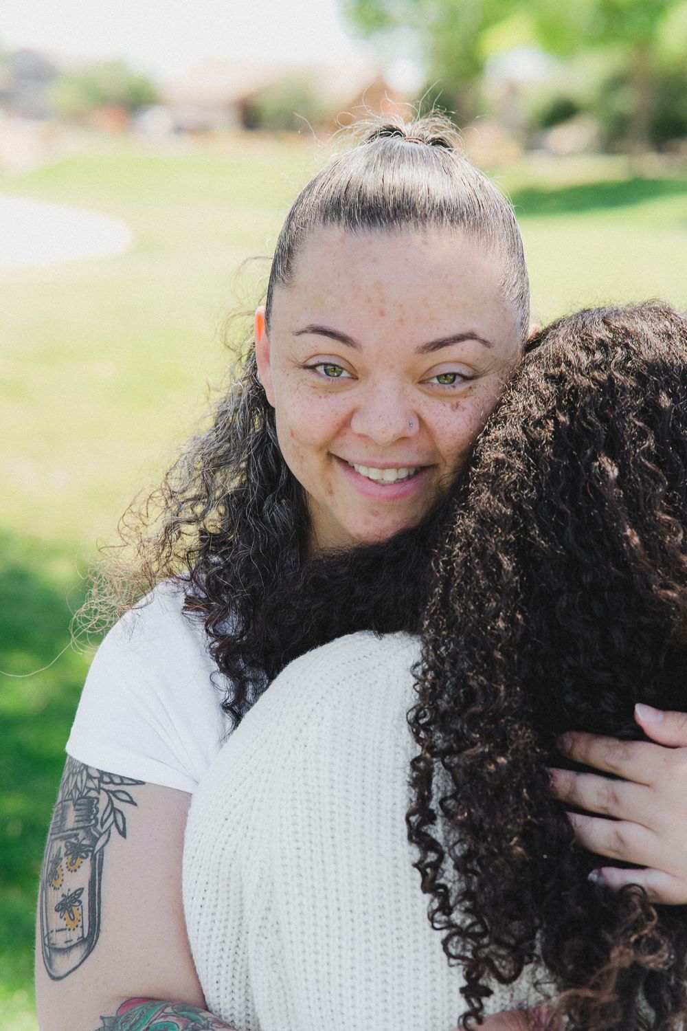 Two people embrace outdoors in a sunny garden setting with natural curly hair and a white shirt visible.
