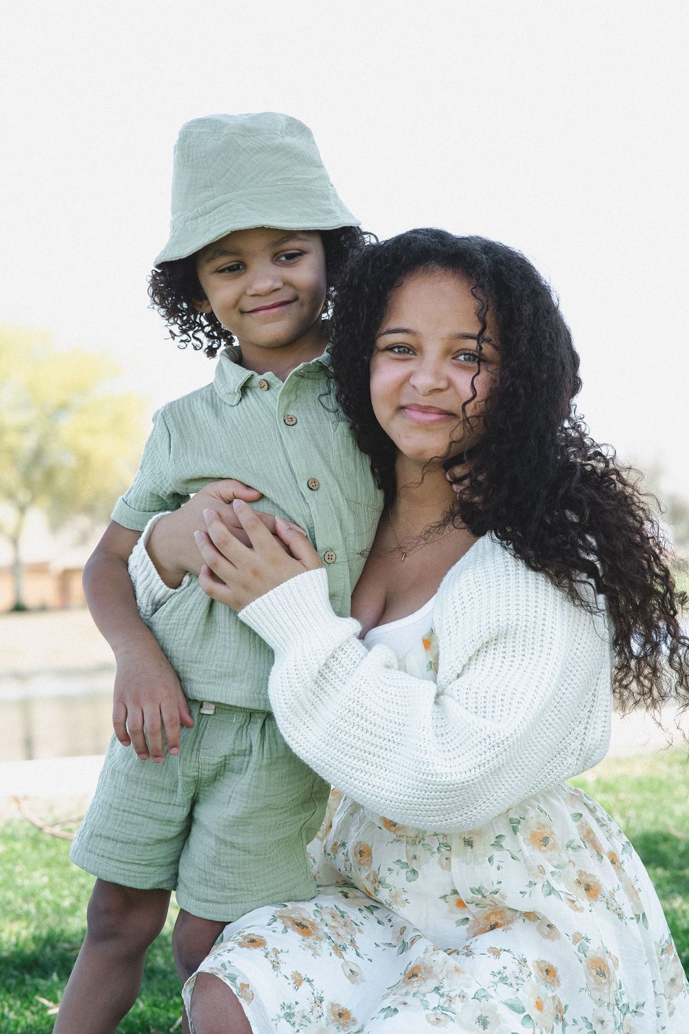 Two people sharing a warm embrace outdoors, wearing a white sweater and mint green outfit with a bucket hat.