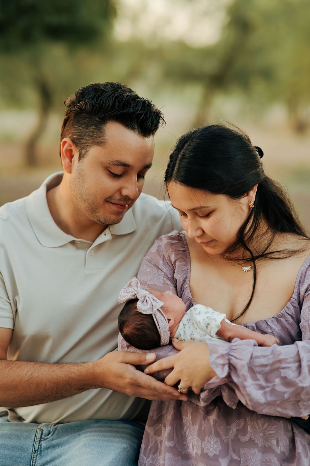 A couple tenderly gazes at their newborn baby outdoors, with the mother in a purple dress and father in a white polo.
