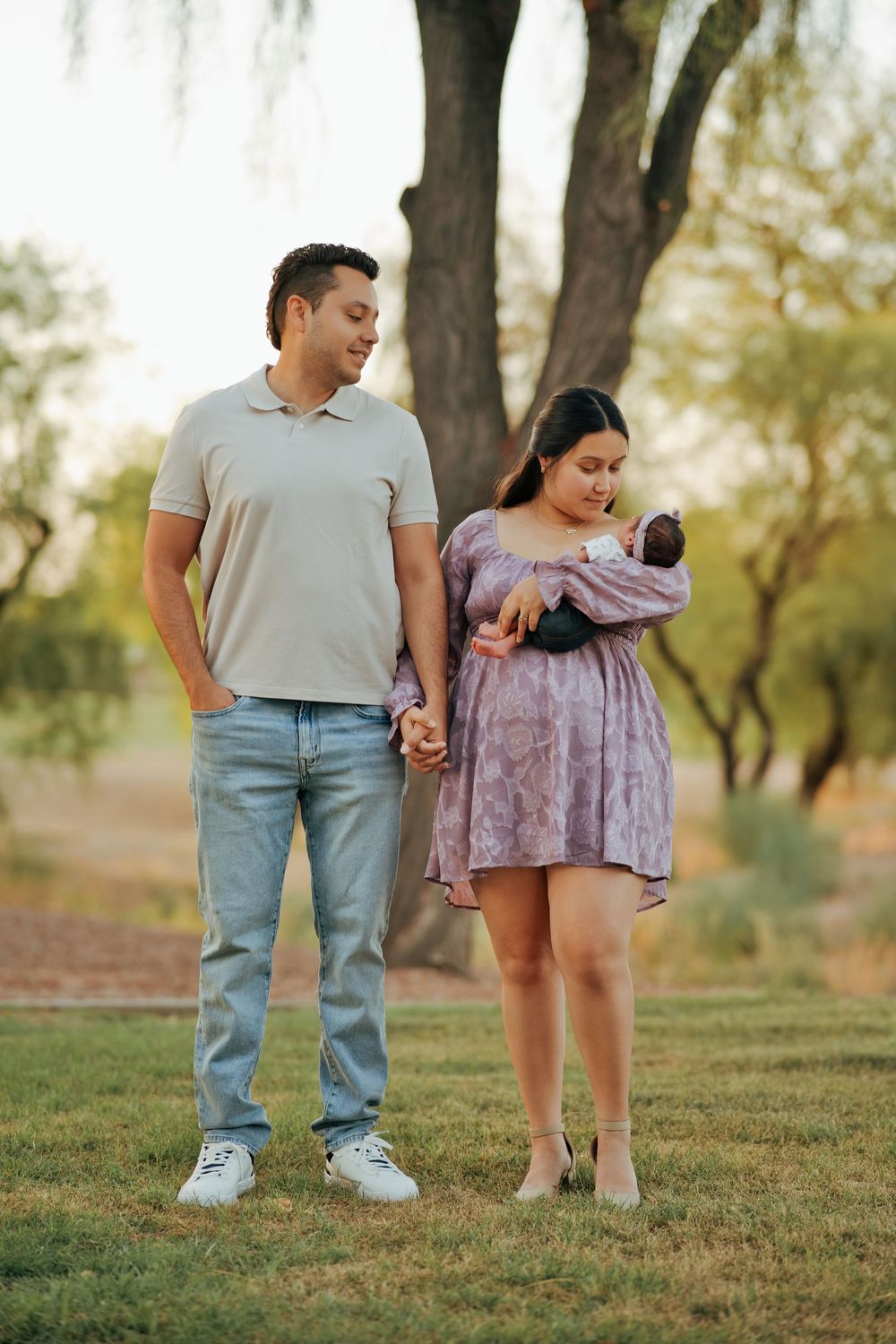 A couple stands outdoors holding hands, with the woman cradling a newborn baby, surrounded by lush green trees.