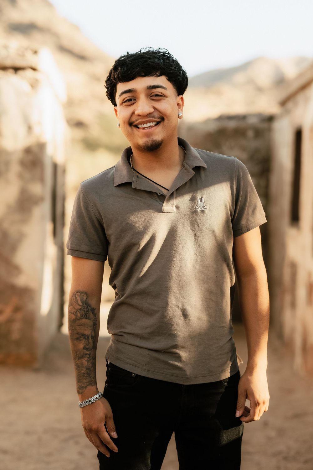 A person wearing a grey polo shirt smiles warmly while standing outdoors near desert buildings in natural lighting.