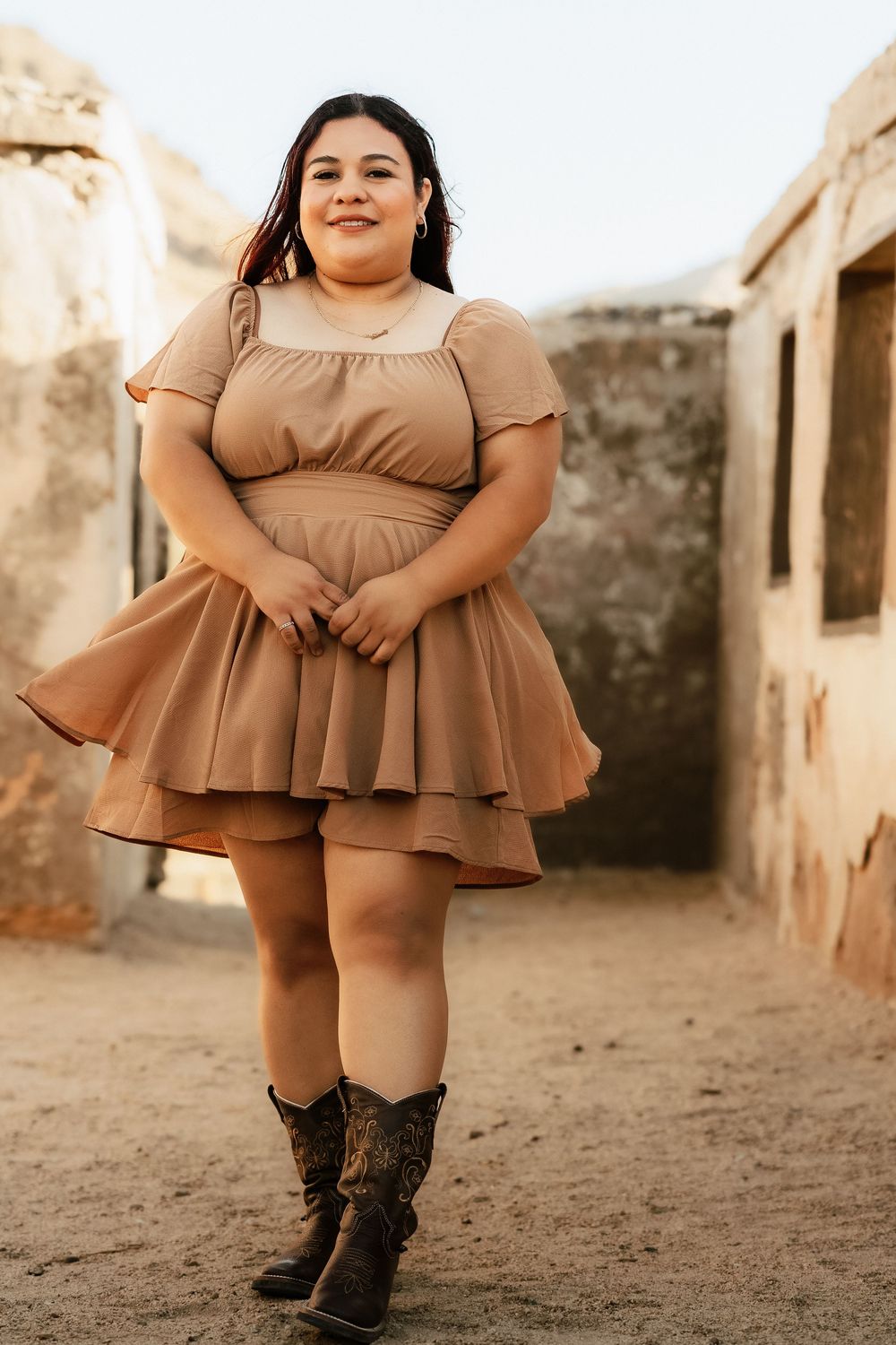 A plus-size model in a brown ruffled dress and cowboy boots poses outdoors in rustic setting during golden hour.