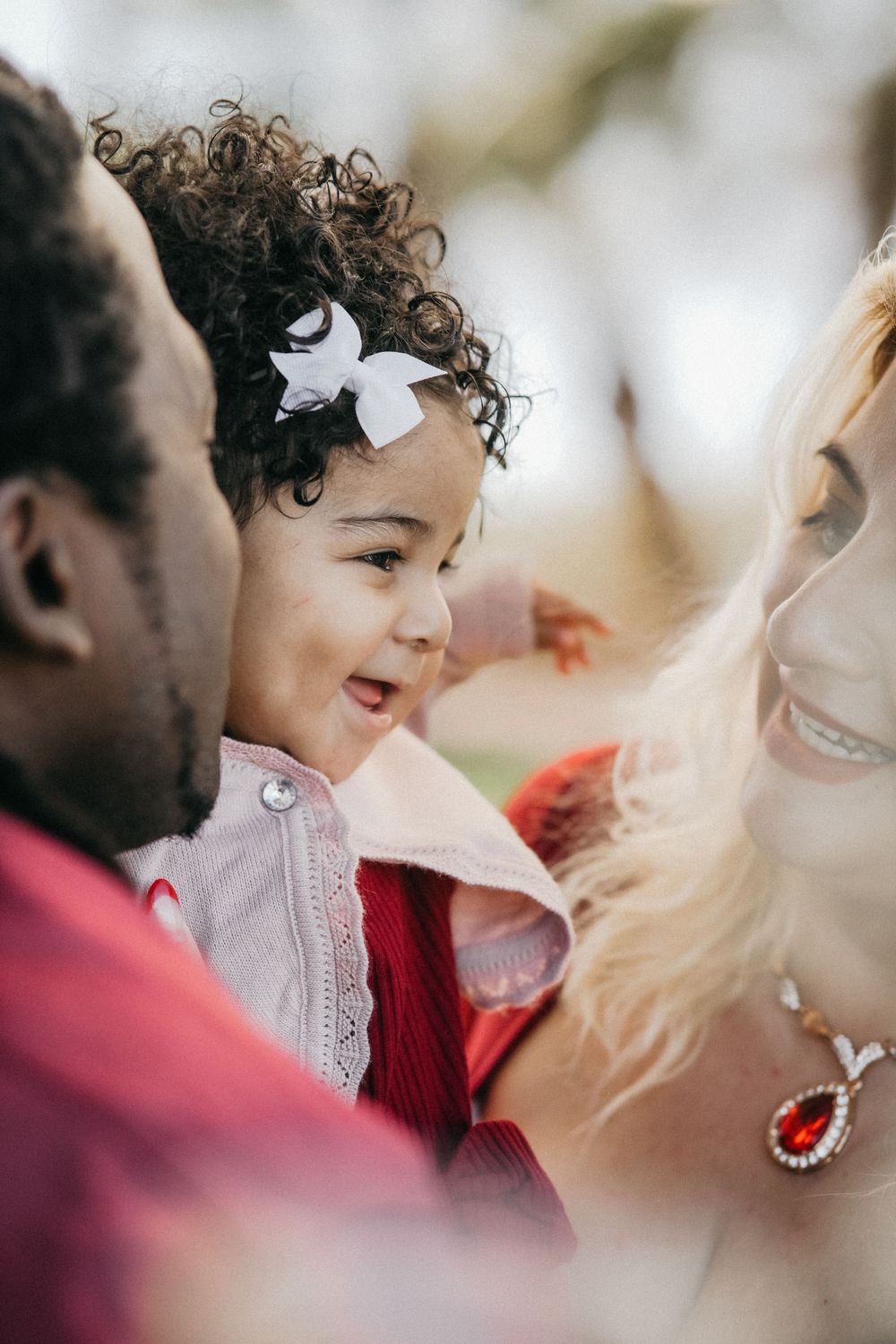 A joyful child with curly hair and white bow wearing a red outfit smiles between two adults at a gathering.