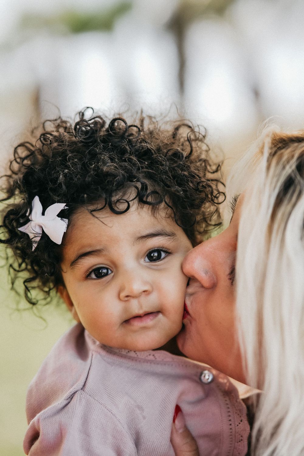 A small child with curly dark hair and a white bow wearing pink gets a gentle kiss from a loved one.