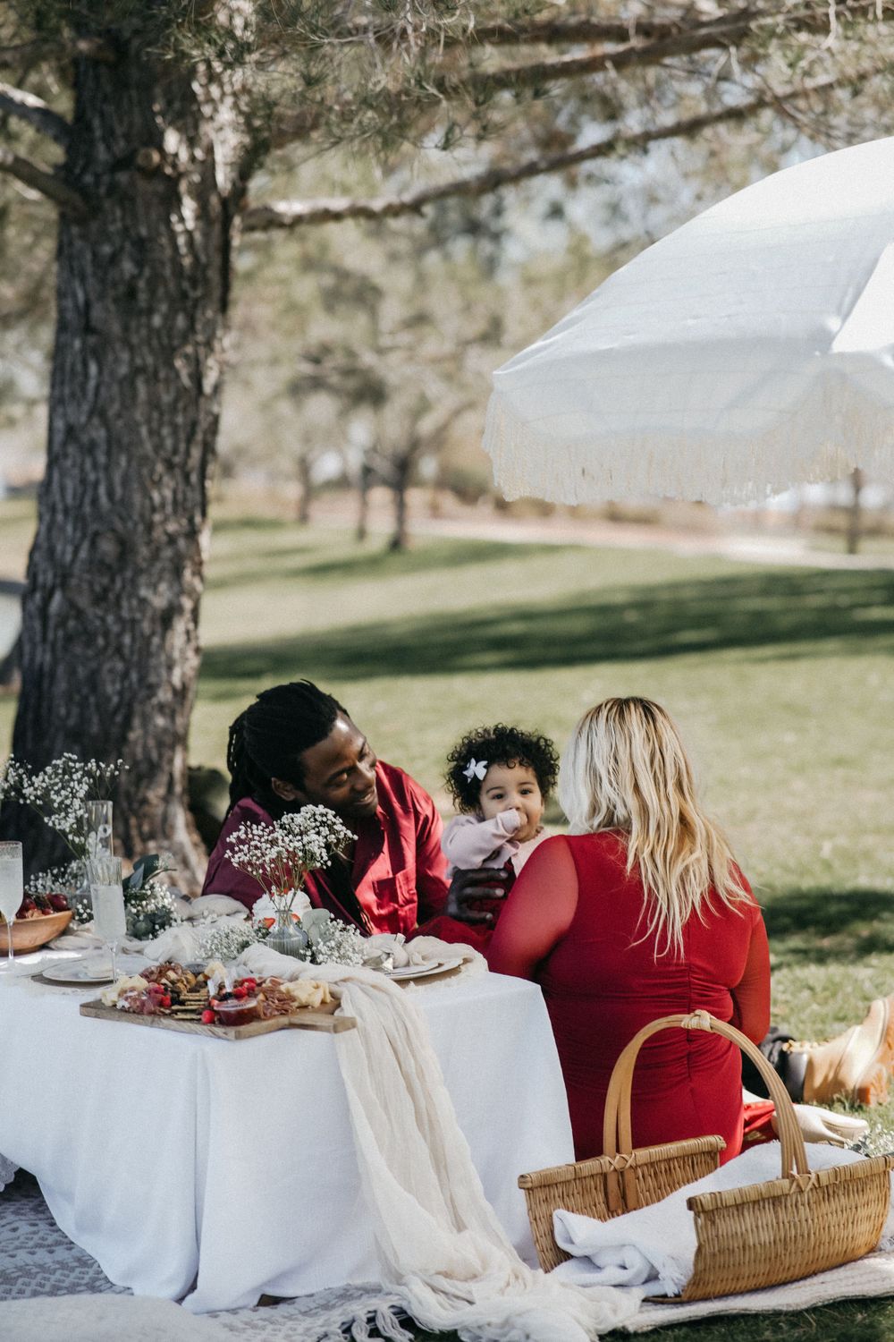 A small group enjoys an outdoor picnic under a white umbrella with a spread of food on a white tablecloth near a tree.