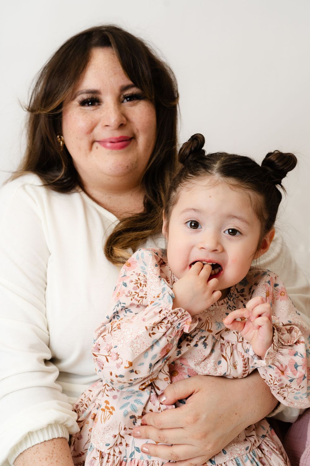 A parent and young child in matching white and floral outfits pose together against a plain backdrop for a portrait.