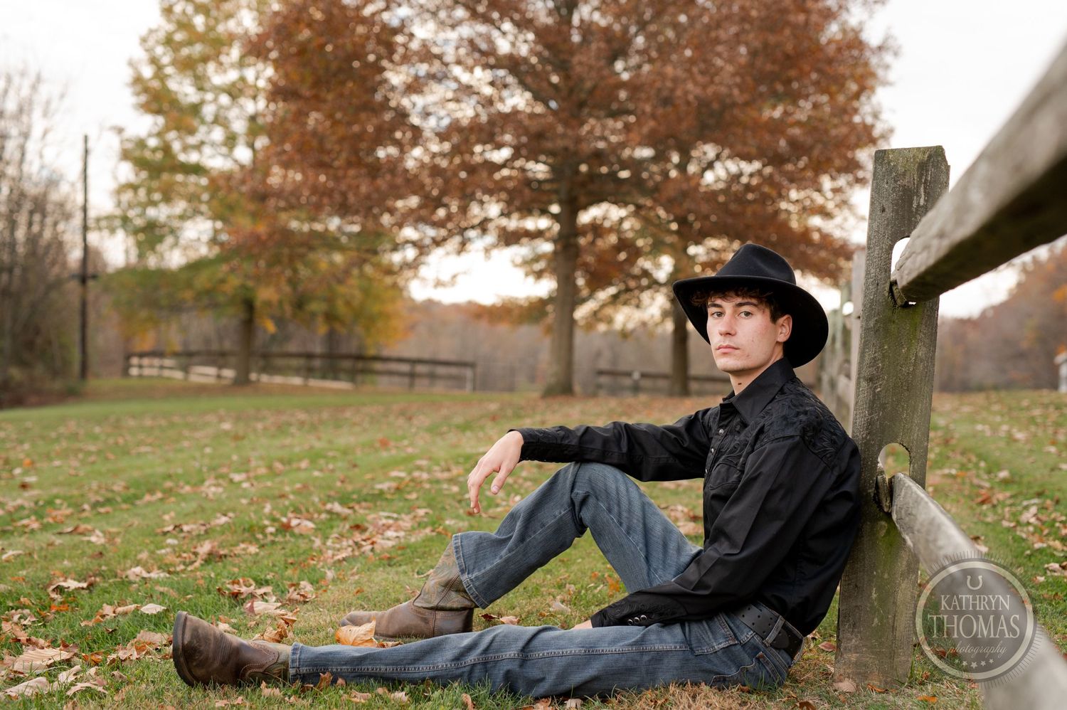 Casual western portrait leaning against a wooden fence surrounded by fallen autumn leaves.