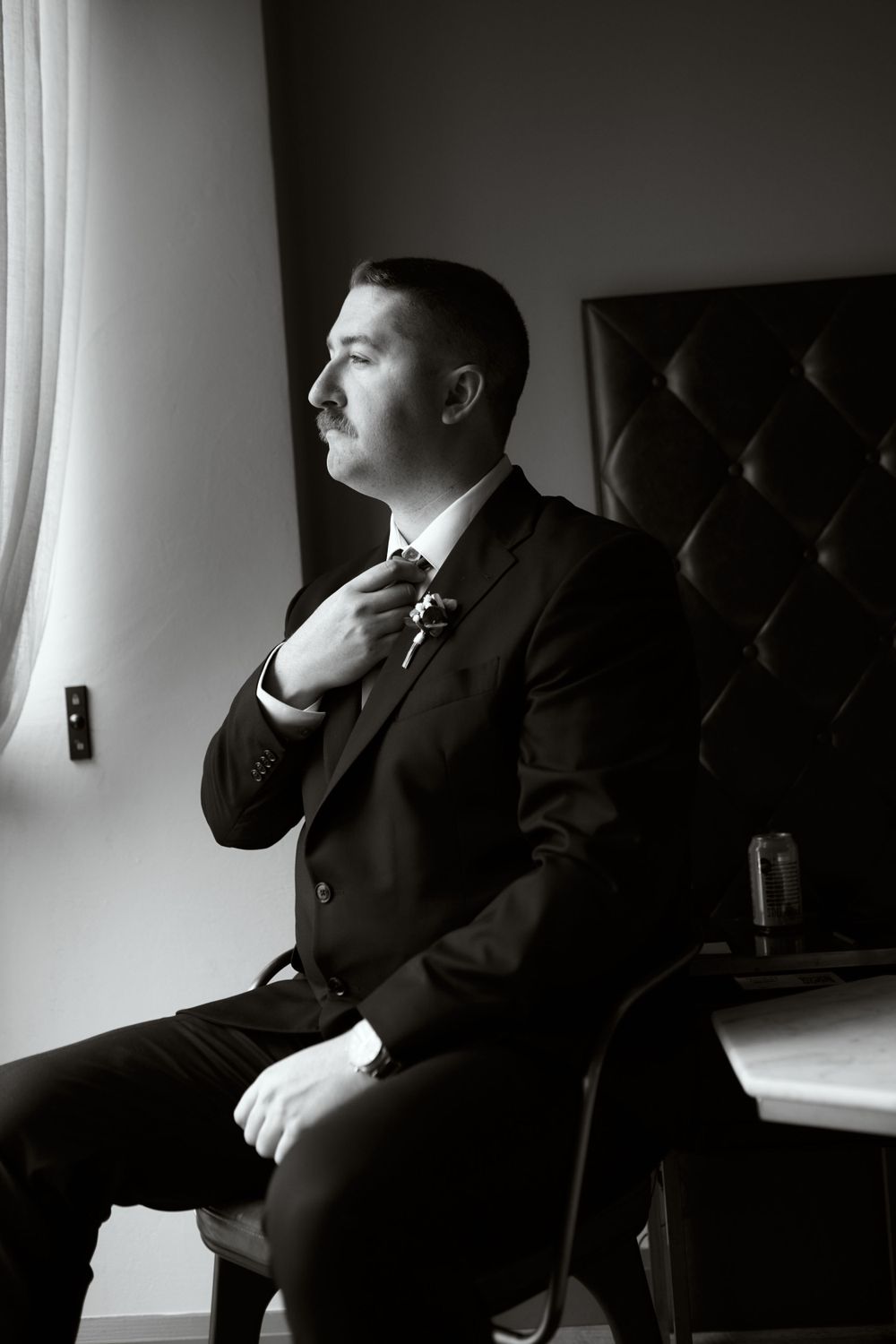 Dramatic black and white portrait of groom adjusting tie by window.