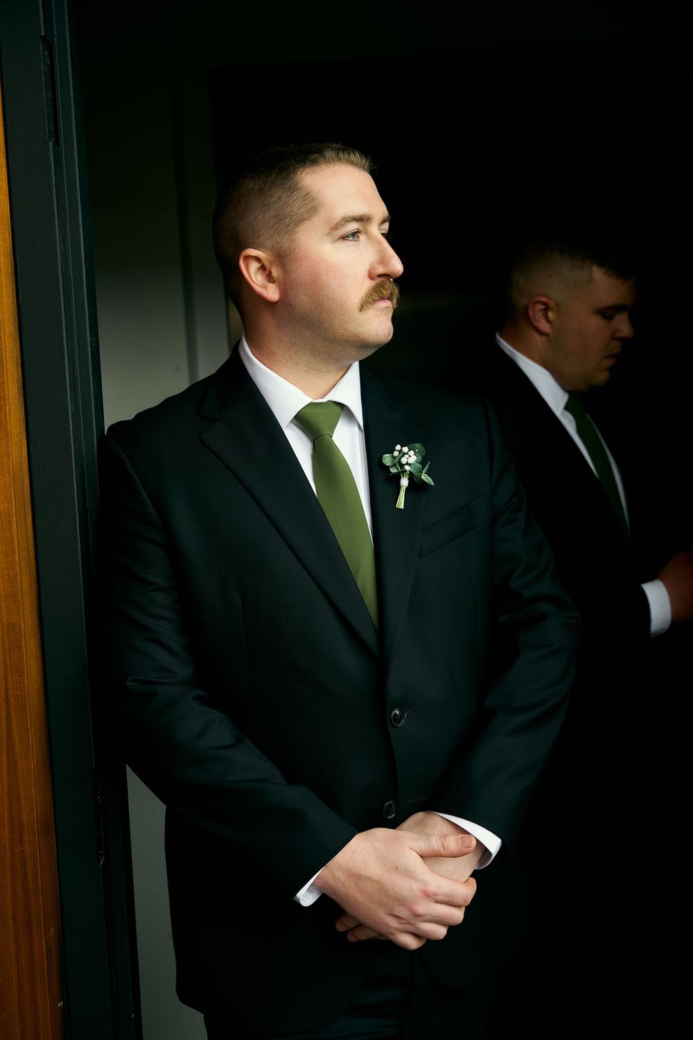 Men in formal black suits with green ties stand together in a dimly lit hallway.