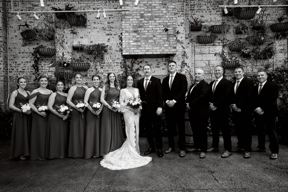 Wedding party poses against rustic brick wall backdrop for formal group photo.