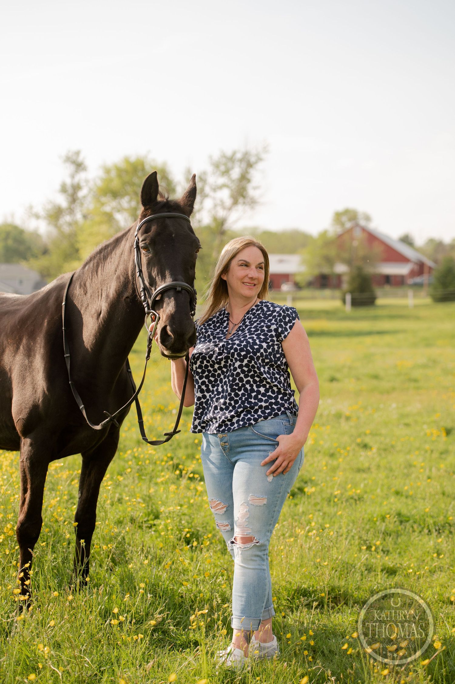 A person in casual wear stands with a black horse in a spring meadow with wildflowers.