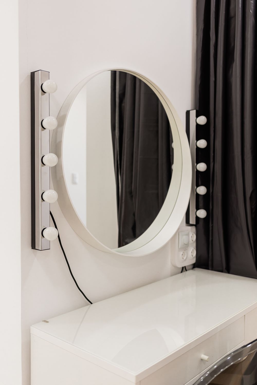 Close-up of a white vanity mirror with LED lights mounted on a white wall next to dark curtains.