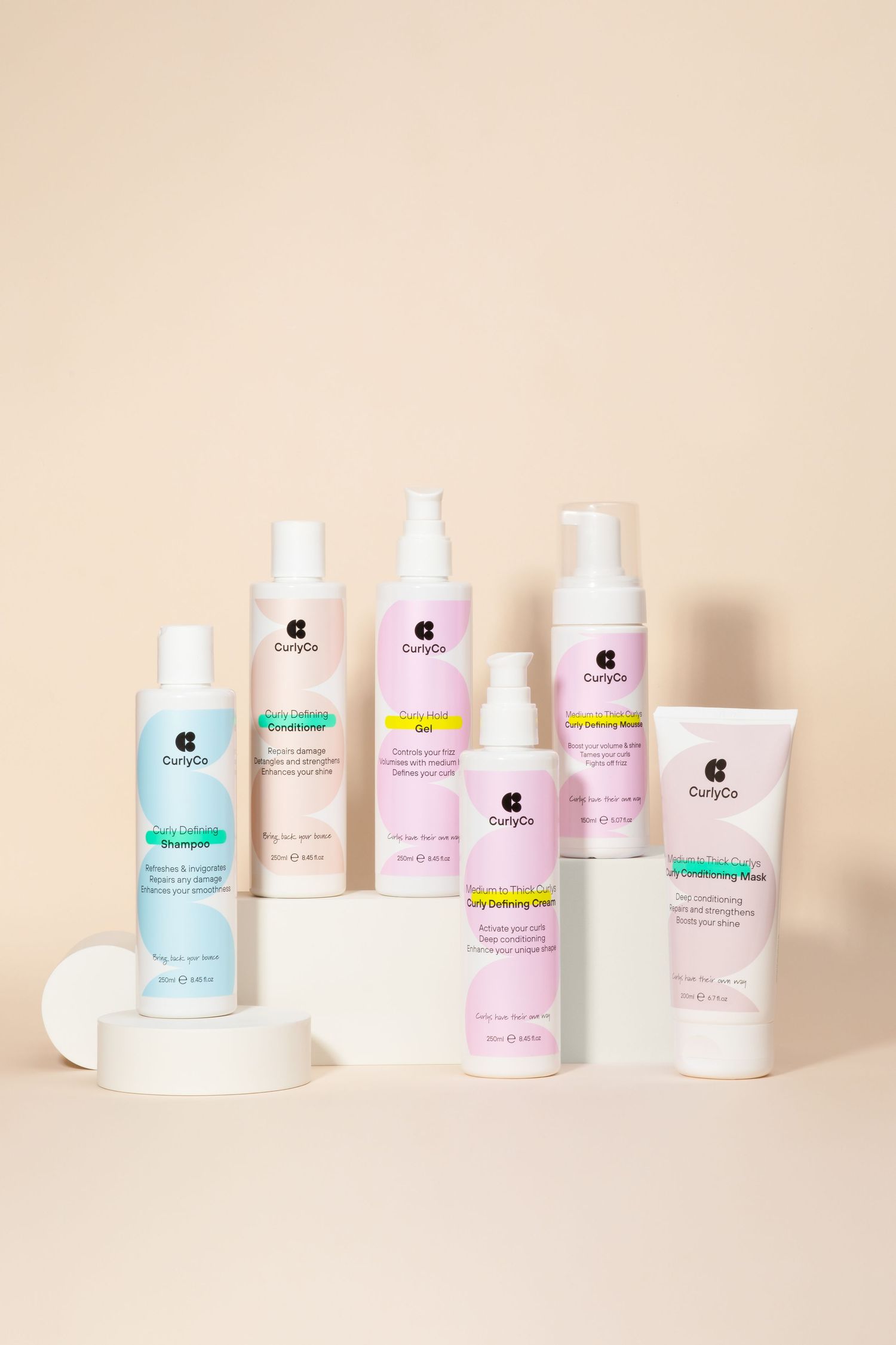 Collection of CurlyGo hair care products displayed on cream coloured background with white pedestals.