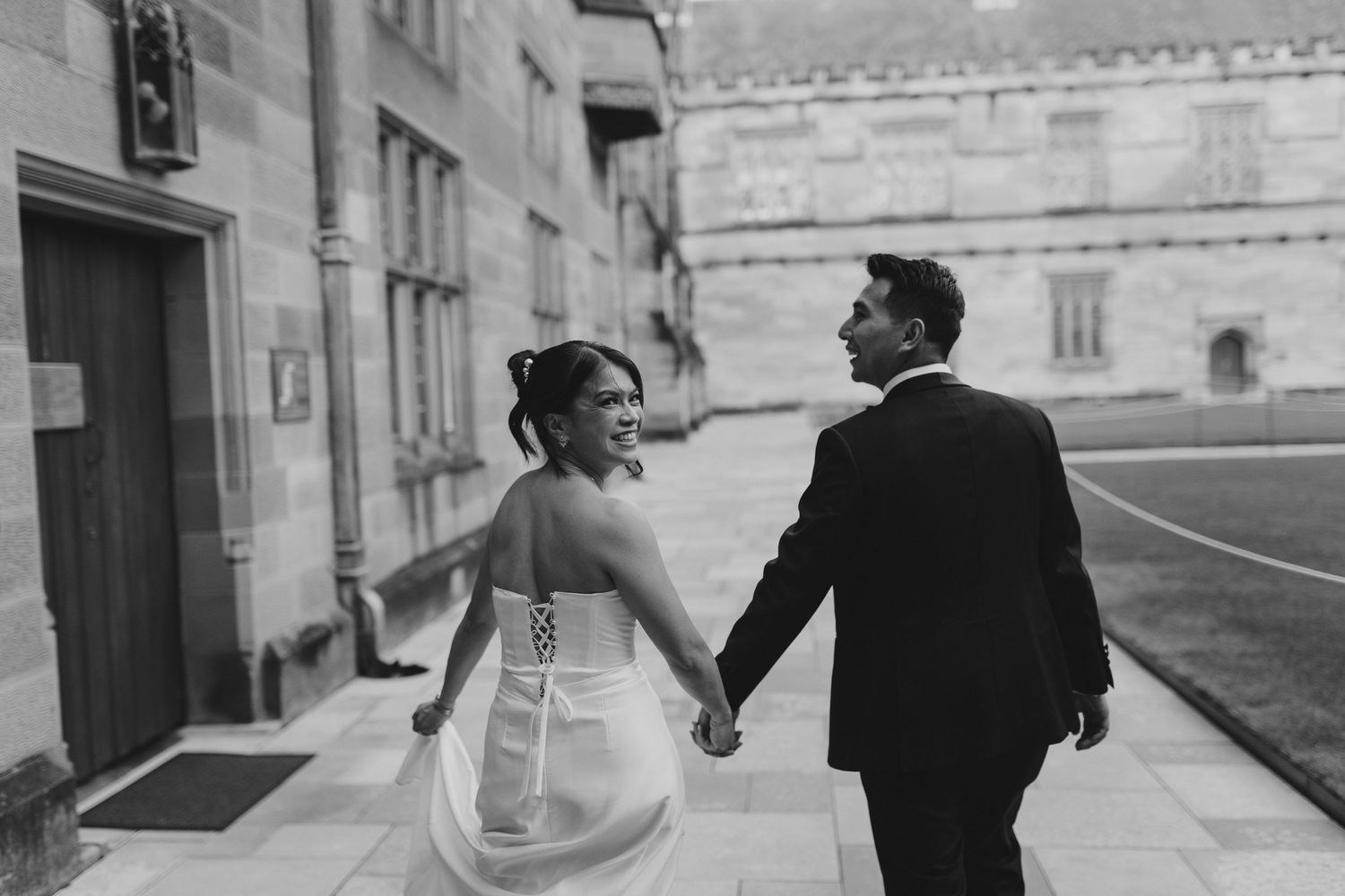 Black and white photo of couple walking hand in hand along historic stone building exterior.