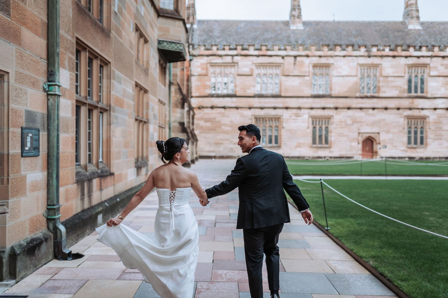 Couple walks along historic university building with green courtyard in background.