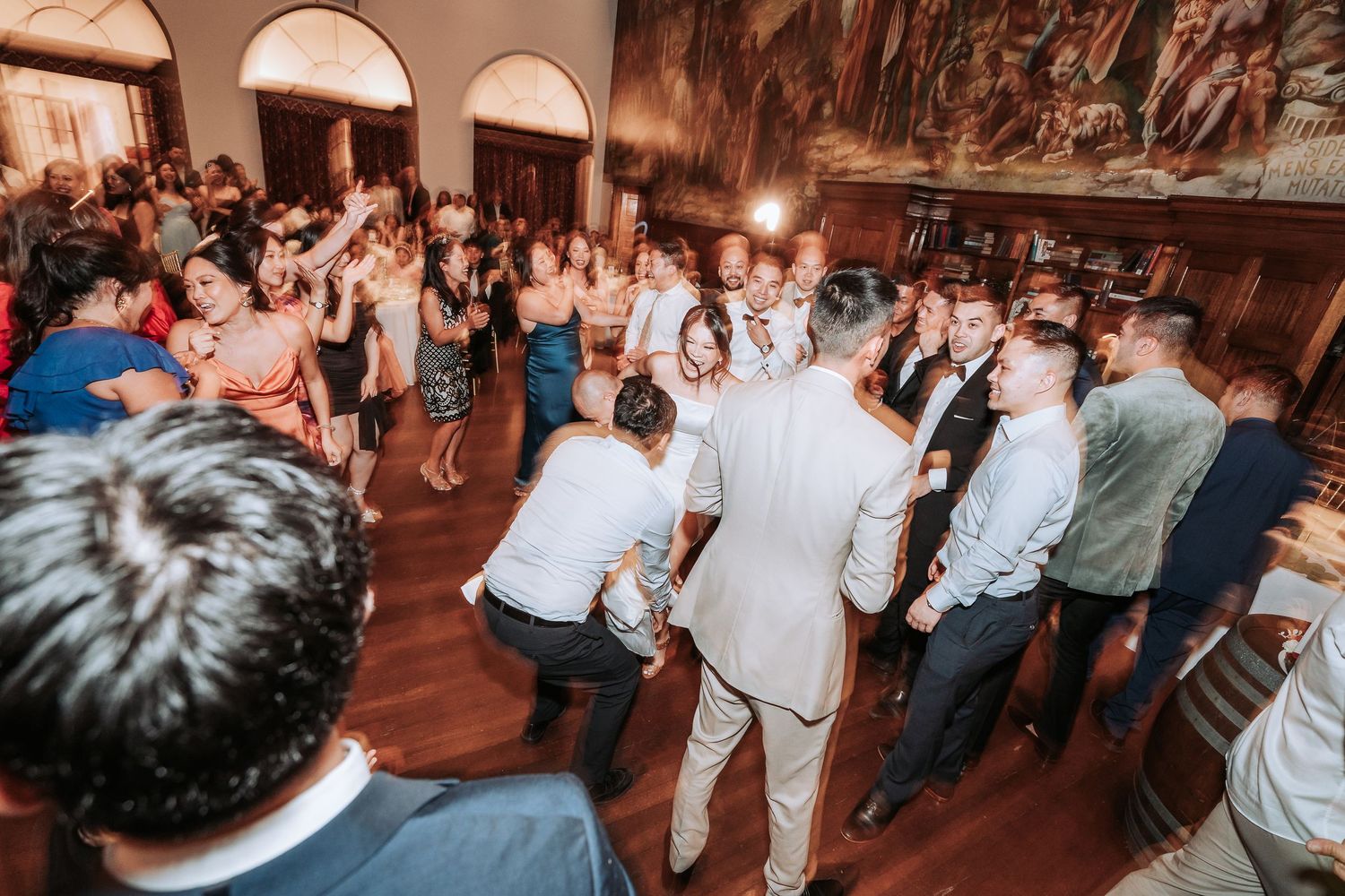 Wedding guests dancing and celebrating on a crowded dance floor in an ornate reception hall.
