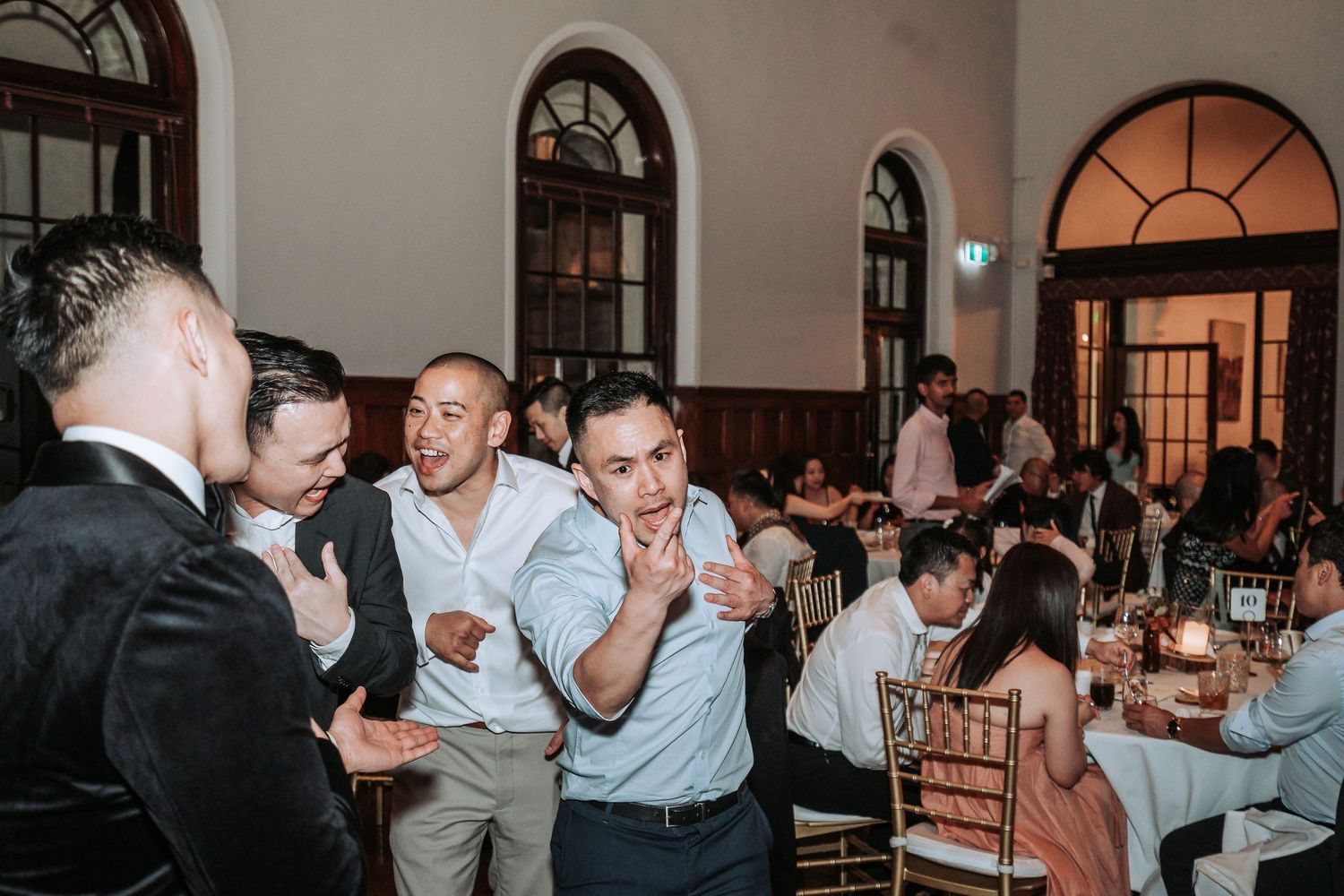 Wedding guests enjoying lively conversation at an elegant reception with arched windows.
