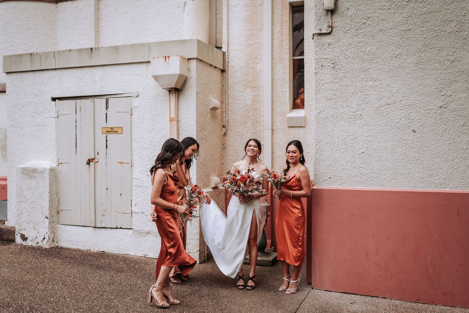 Wedding party in rust-colored dresses poses against a white and red painted building wall.