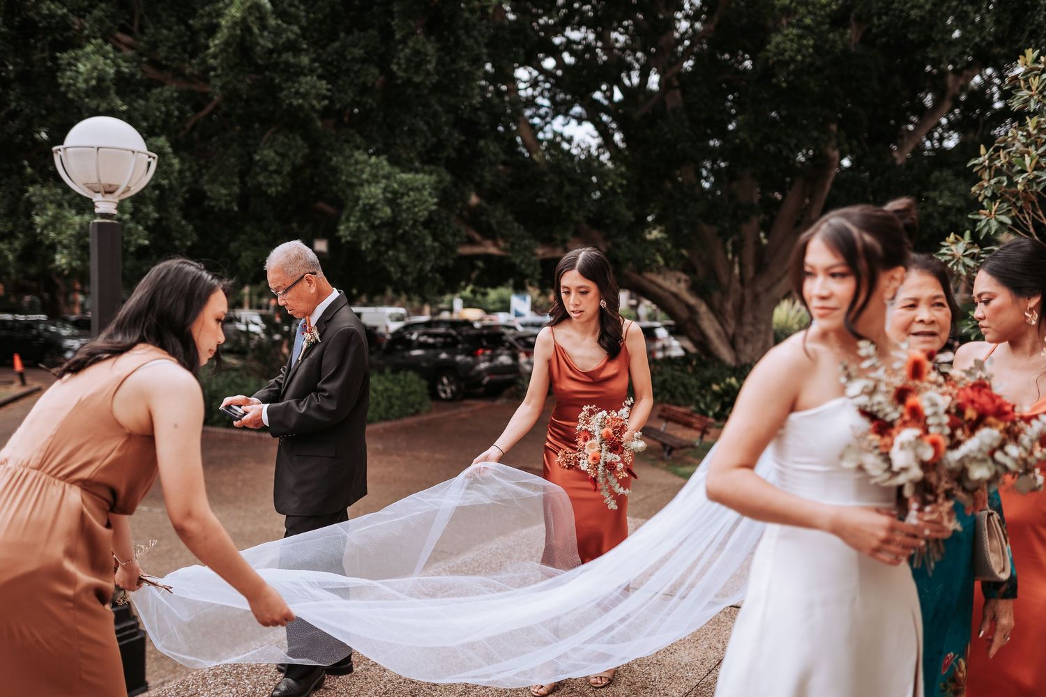 Bridesmaids in terracotta dresses help adjust the bride's flowing veil outdoors.