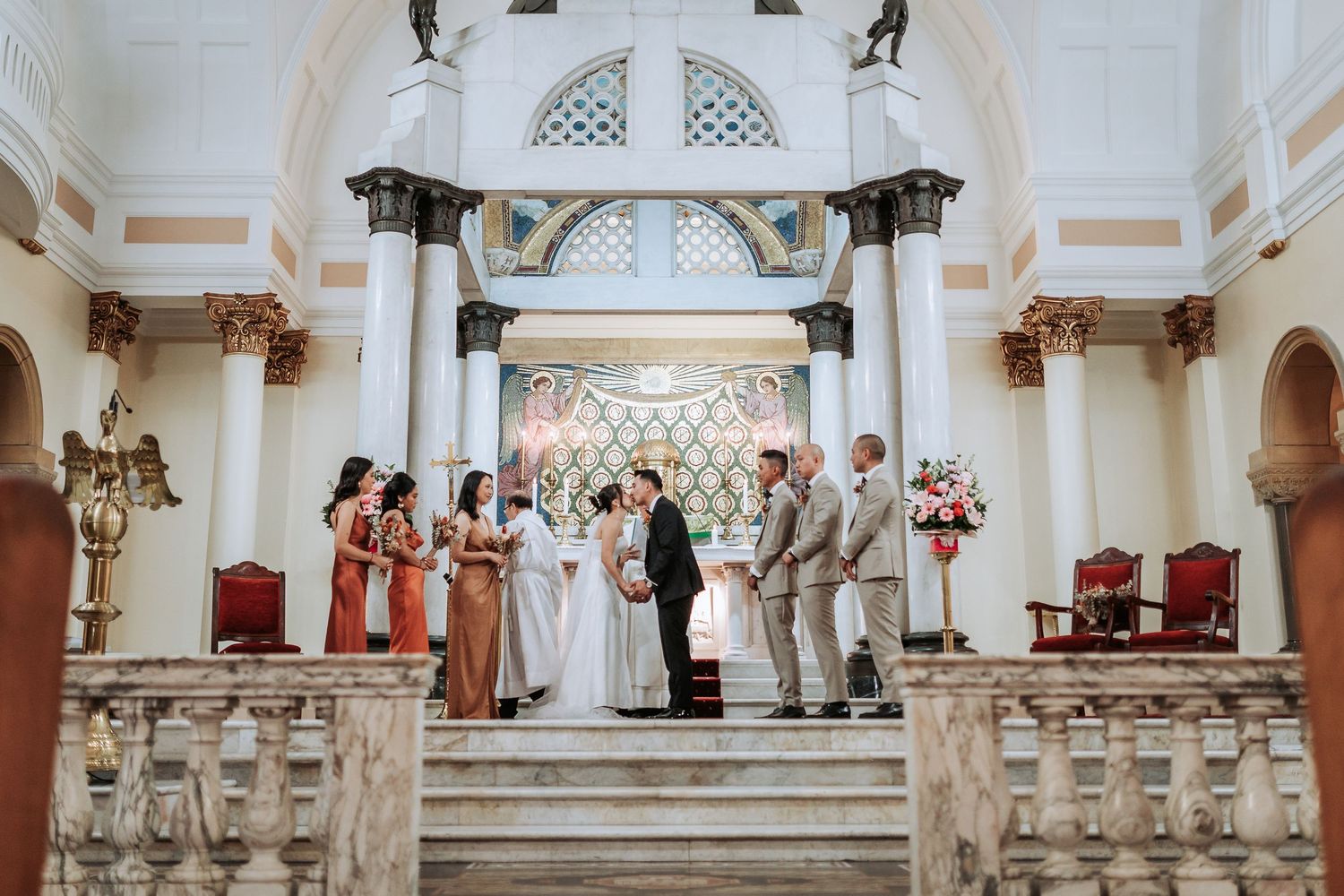 Wedding ceremony in a grand white chapel with coral-colored bridesmaids dresses and elegant architecture.