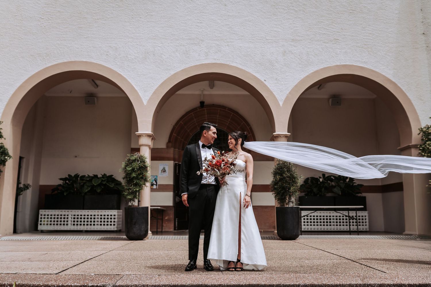 Newlyweds pose under arched columns while the bride's veil flows in the wind.