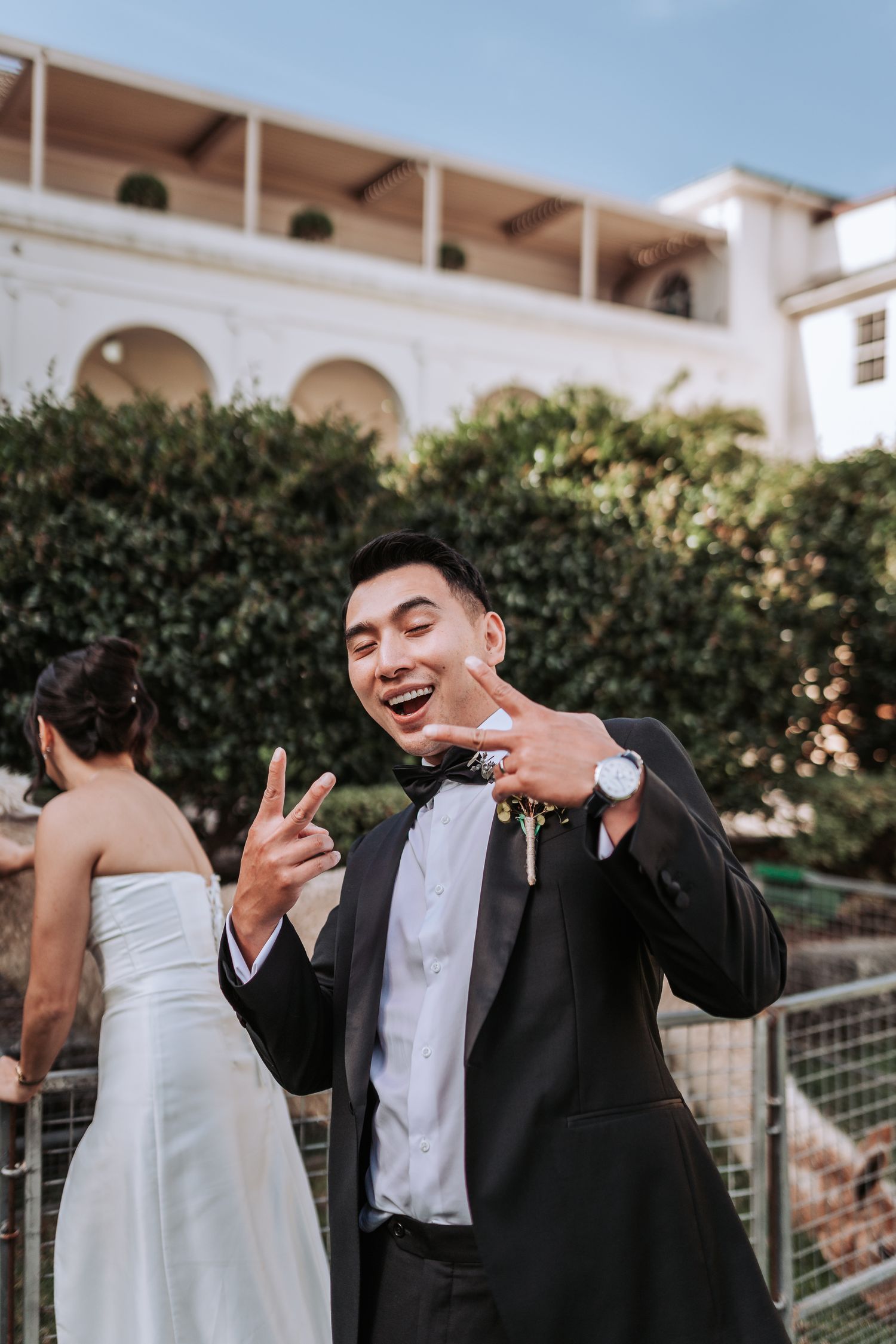 Wedding party member in tuxedo makes playful gestures at outdoor venue with arched architecture.