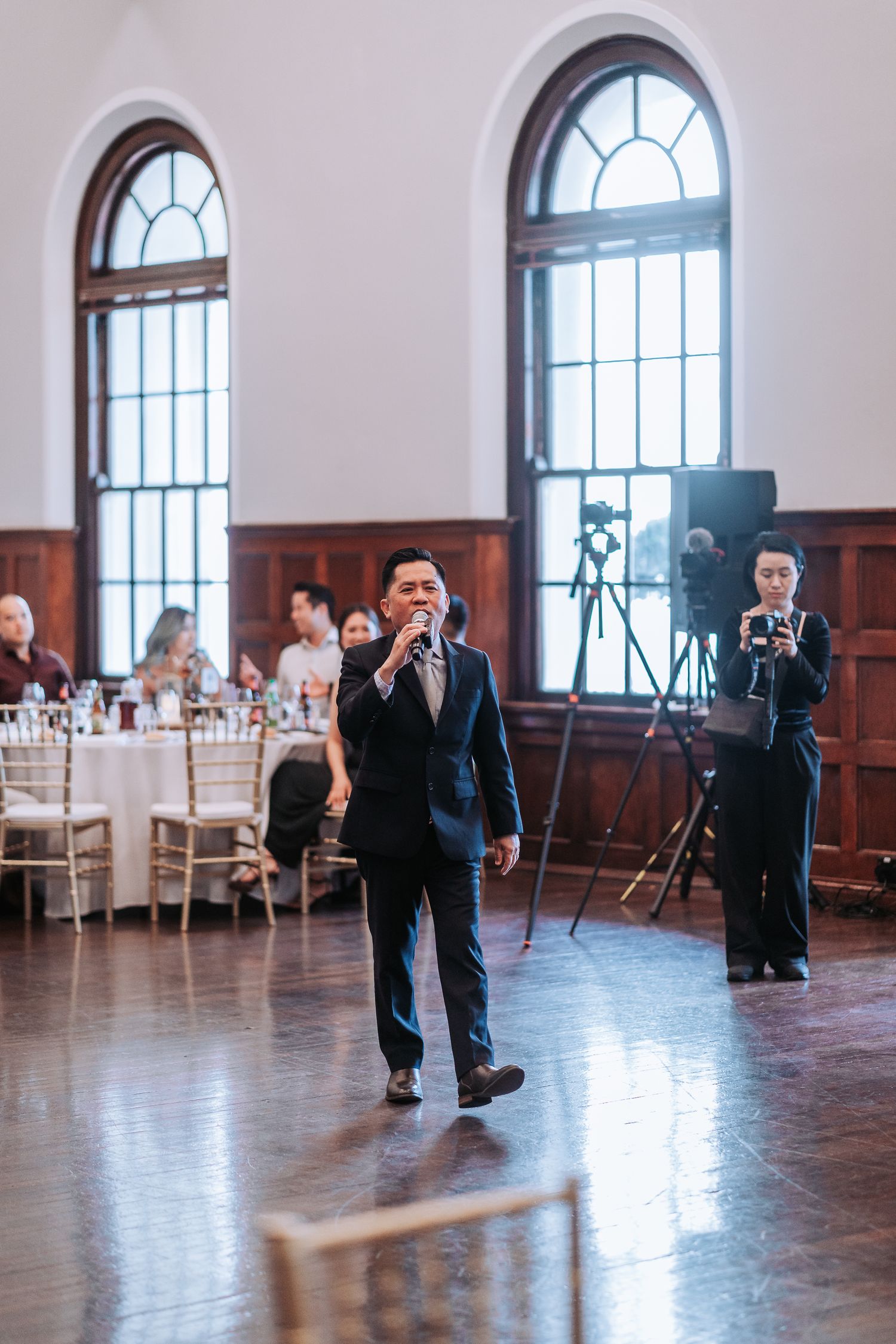 Person speaking into microphone in formal attire inside an elegant venue with arched windows.