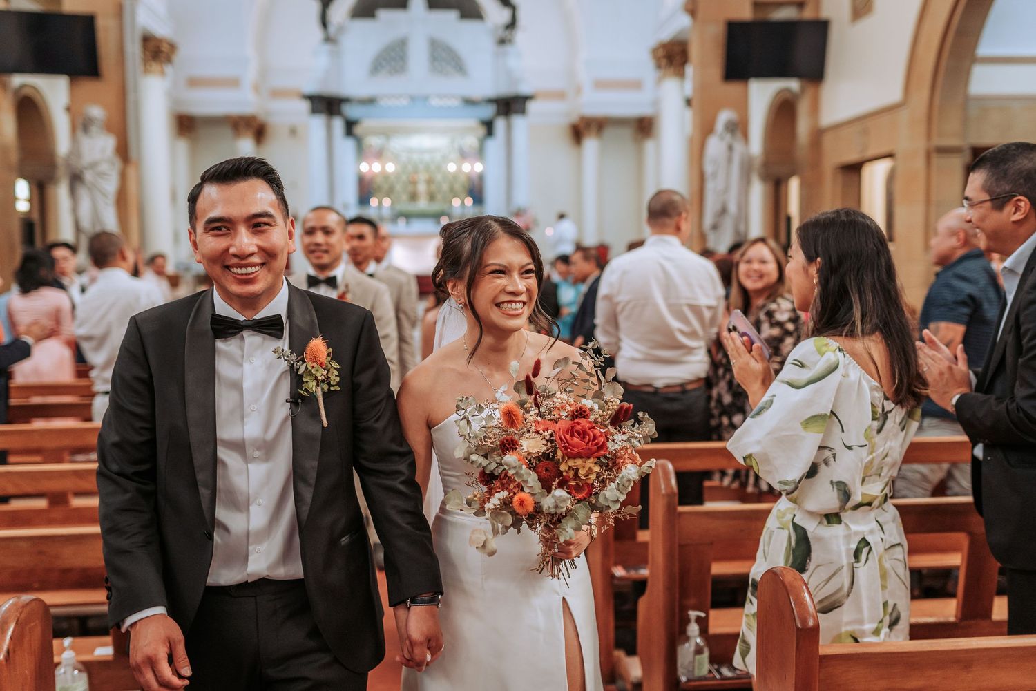 Newlyweds walk down the church aisle smiling while guests look on from wooden pews.