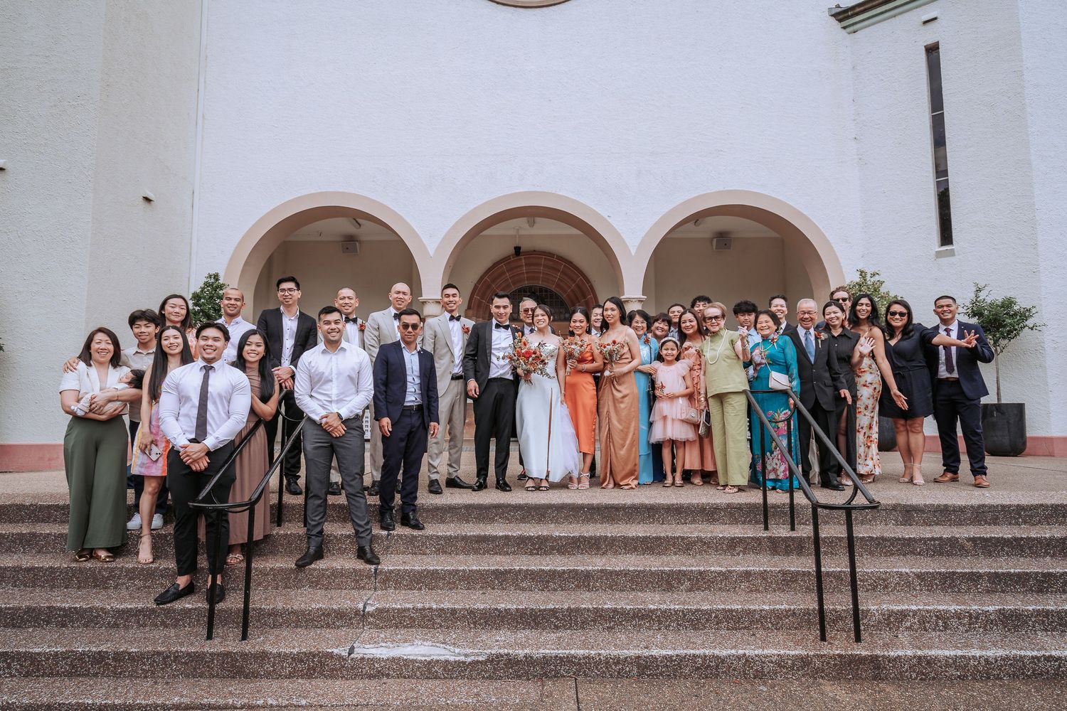 Large wedding party group photo on church steps with diverse formal attire.