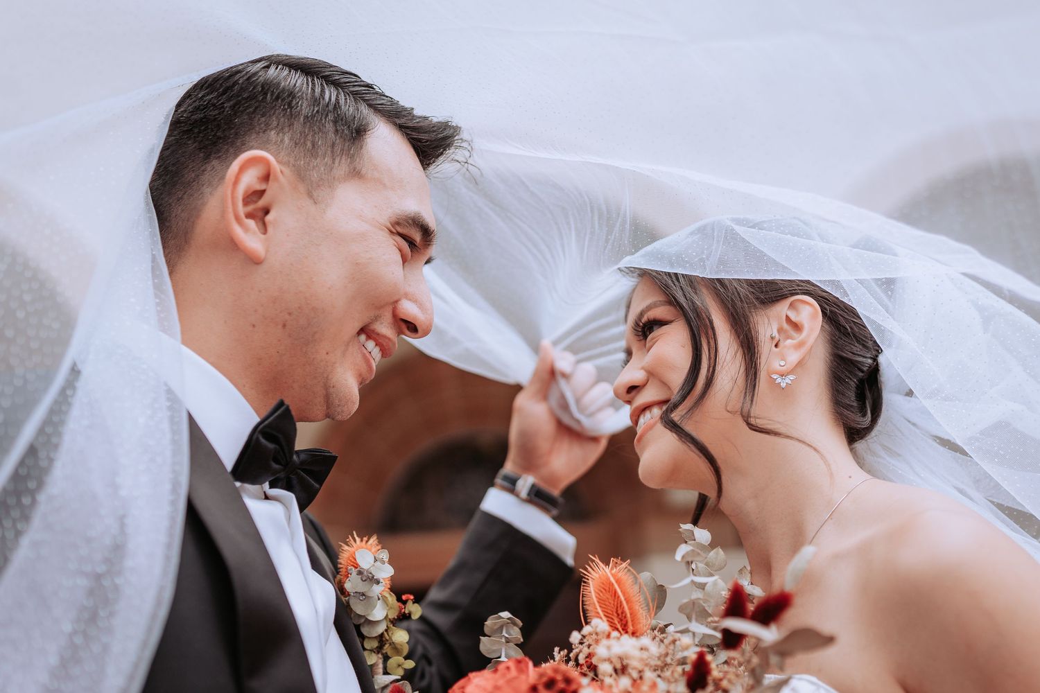 Intimate moment between newlyweds under wedding veil with coral flowers in foreground.