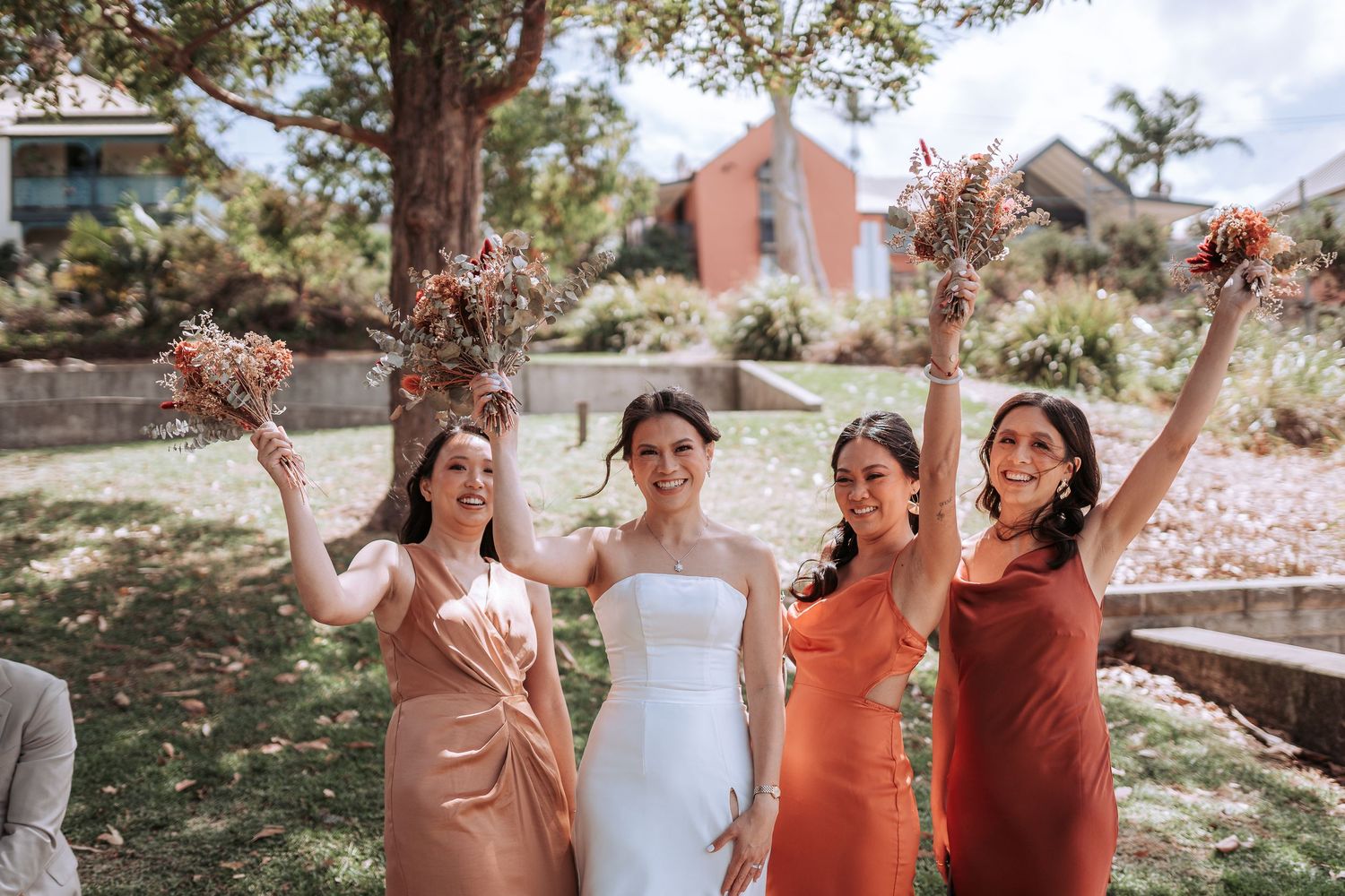 A bridal party in autumn-toned dresses celebrating joyfully on a lawn with a brick building in the background.