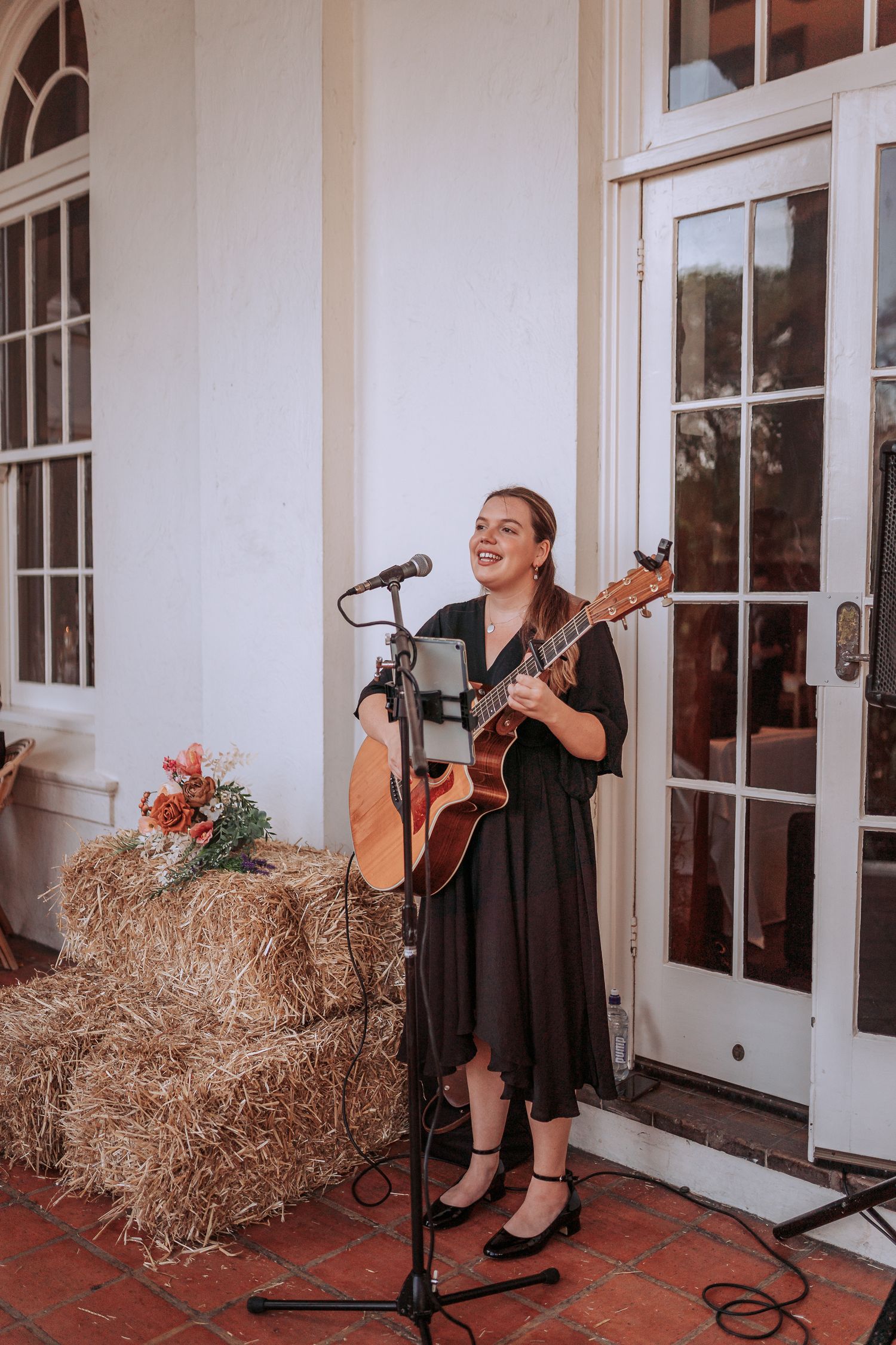 A musician in a black dress performs with an acoustic guitar on a porch with hay bales and french doors.
