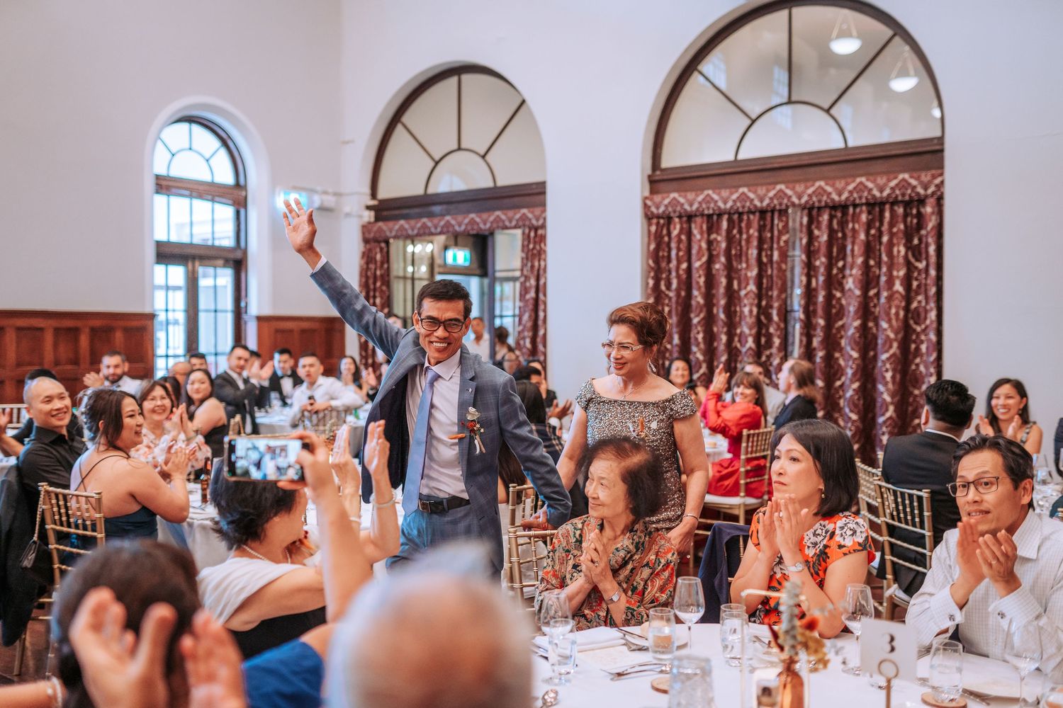 Wedding reception guests cheering and raising glasses in an elegant dining room with arched windows.