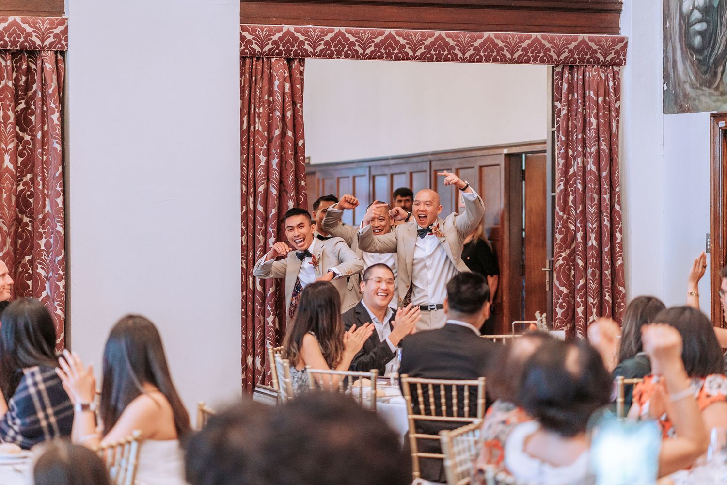 Wedding guests celebrating and waving napkins in an elegant reception hall with arched windows.