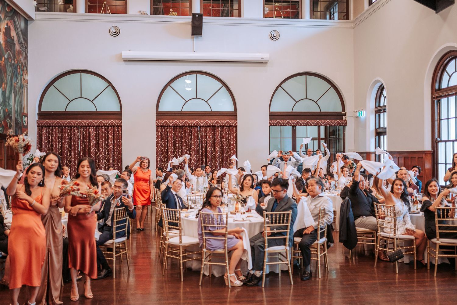 Wedding reception in a grand hall with coral-colored curtains and arched windows as guests celebrate.