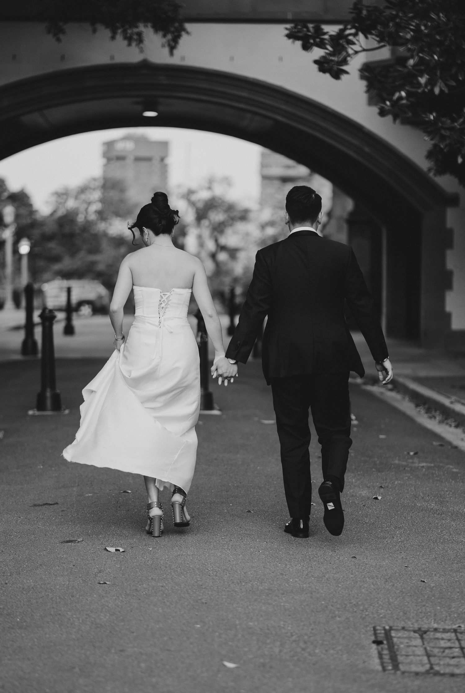 Black and white photo of newlyweds walking hand in hand under an archway.