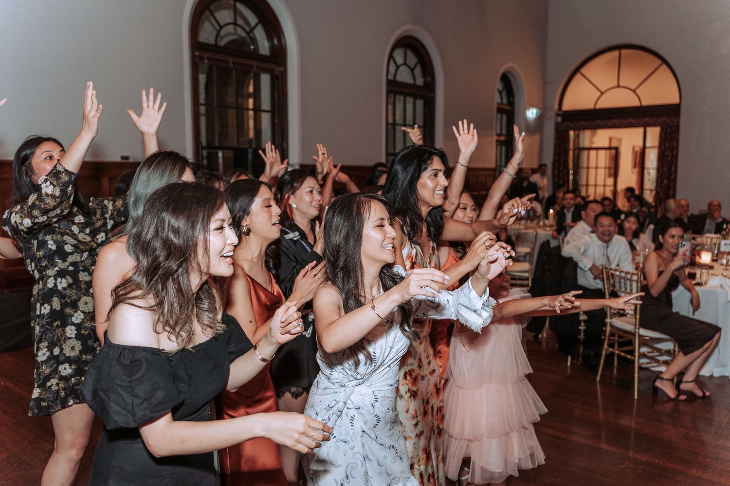 Wedding guests dance with raised arms in elegant reception hall with arched windows.